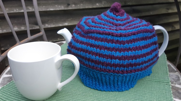 White mug and tea pot on green placemat. Tea pot has a blue and purple striped knit cozy. 