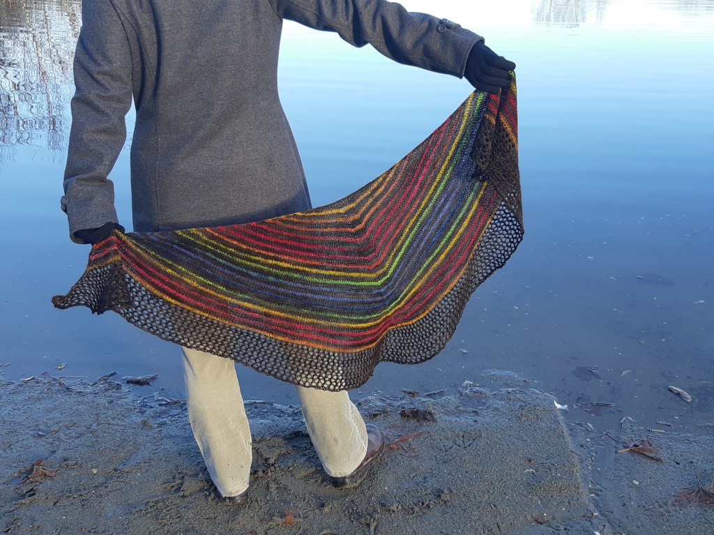Woman with back to camera wearing a grey wool coat and holding a striped heart-shaped shawl (brown and rainbow stripes) in front of a lake. 