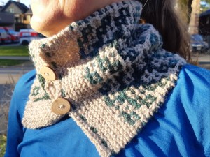 Smiling woman in blue shirt wearing a handknit cowl in cream and blue/green yarn in a garter mosaic structure with two wood buttons. 